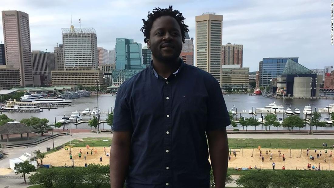 A Black man is posing for a photo before a sprawling cityscape behind him.
