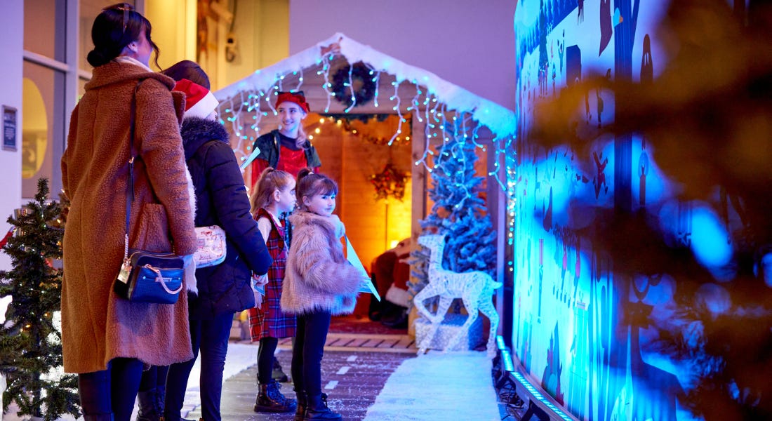 Four people - two adult and two children - queuing up outside a wooden Santa's grotto, standing on a snowy path