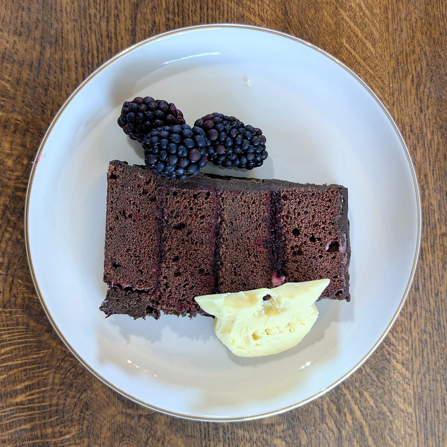 A tall slice of chocolate layer cake sits on a white plate with a gold rim. Thin layers of dark sponge alternate with smooth filling, forming clean, even stripes. Three glossy blackberries rest on top, and beside the slice lies a small broken piece of the white chocolate skull decoration. The plate sits on a wooden surface, its warm grain adding to the cosy, indulgent feel.
