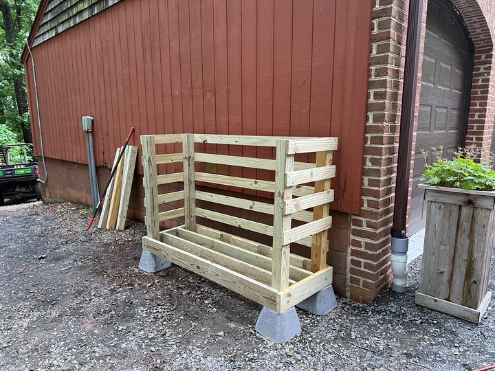 Two side-by-side photos showing a homemade firewood rack before and after being filled. The left image shows a newly built wooden storage rack mounted on concrete deck blocks beside a red barn wall. The right image shows the completed rack packed with neatly stacked split firewood, with additional stacks extending beyond the rack. The photos illustrate the transformation from an empty firewood storage structure to a fully stocked woodshed.