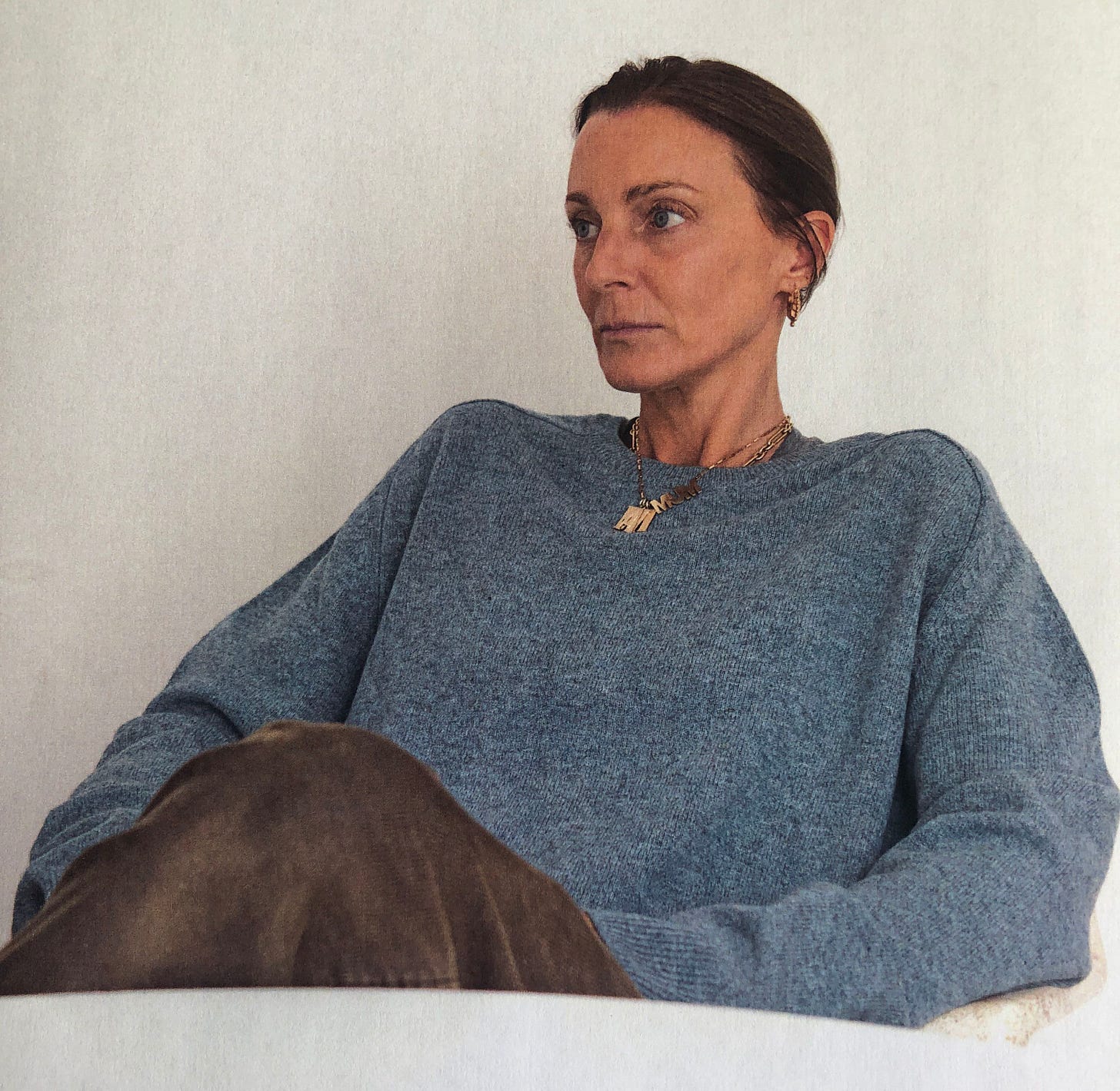 A woman sits behind a white table with her short brown hair pulled back and wearing an oversied gray sweater and brown pants.