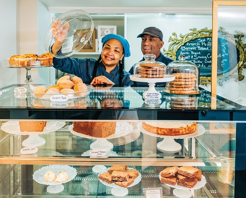 A photo of two bakers from Mid Town Bakers standing behind their case full of baked goods. 