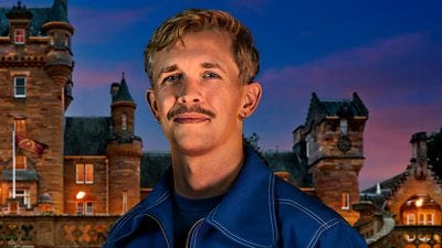 A young man with a moustache smiles to camera, wearing a dark blue jacket with white trim and a dark blue top. He's set against the backdrop of a castle by night. A young man with a moustache smiles to camera, wearing a dark blue jacket with white trim and a dark blue top. He's set against the backdrop of a castle by night.