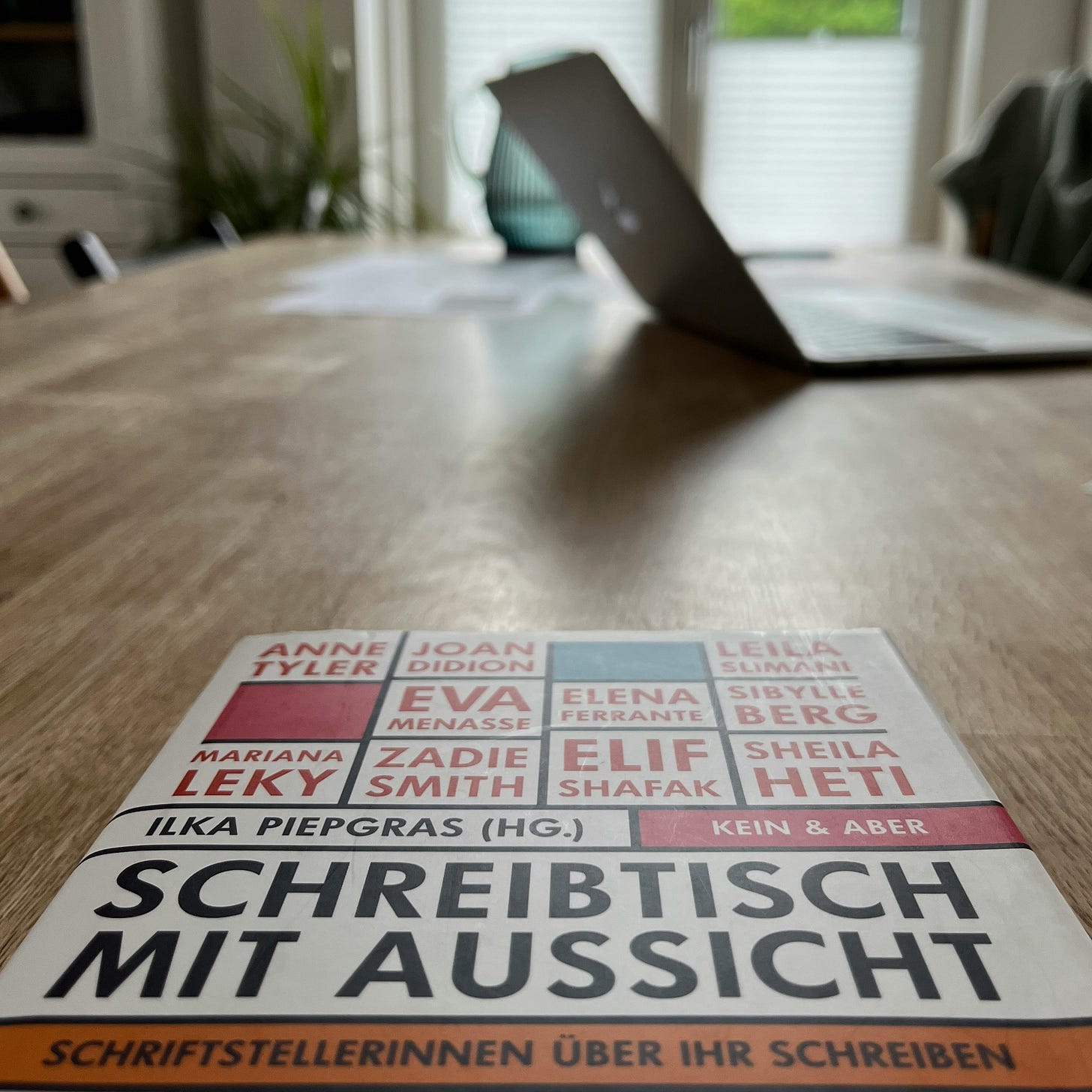 Vorne auf dem Tisch liegt das Buch "Schreibtisch mit Aussicht". Von dort aus blickt man über den gesamten Tisch. Weiter hinten, verschwommen, steht der Laptop und ein Krug mit Wasser. Vorne auf dem Tisch liegt das Buch "Schreibtisch mit Aussicht". Von dort aus blickt man über den gesamten Tisch. Weiter hinten, verschwommen, steht der Laptop und ein Krug mit Wasser.