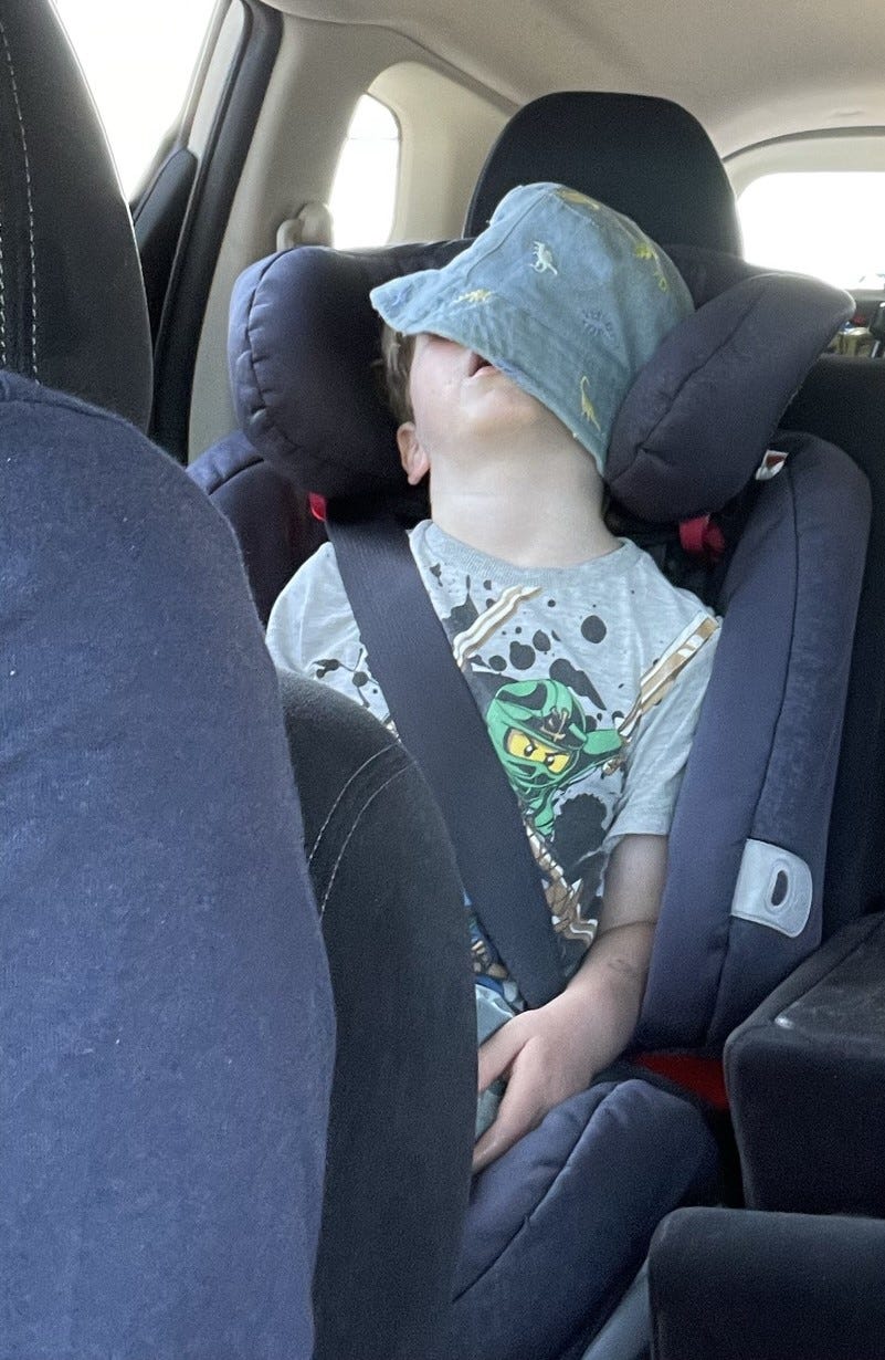 Inside a car with grey seats, a young boy is asleep in his car seat with his denim bucket hat covering most of his face.  His head is tilted back and mouth slightly open.  He is wearing a grey tshirt with lego cartoon on the front. His hand is resting flopped by his thighs.