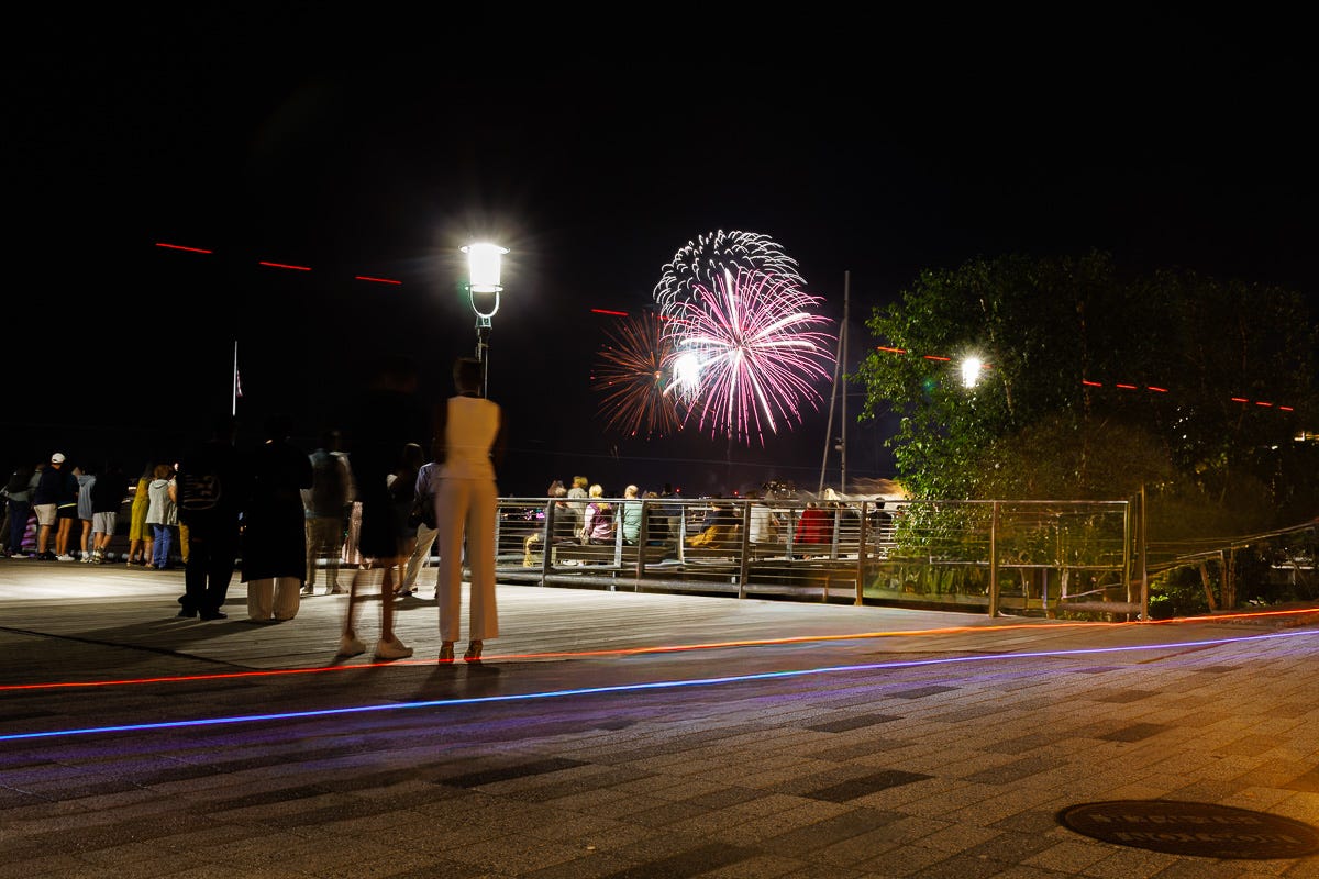 Red and white fireworks over Boston Harbor with streaks of colored light across the pier foreground
