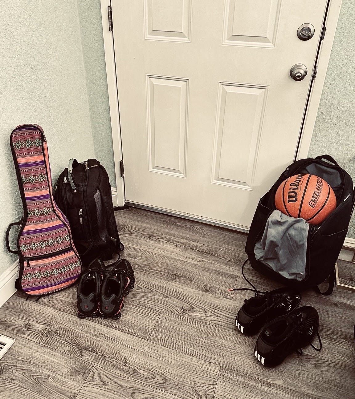 Two backpacks set by a front door: one on the left with a colorful patterned ukulele gig bag and black sneakers, and one on the right with a basketball poking out and high-top athletic shoes.
