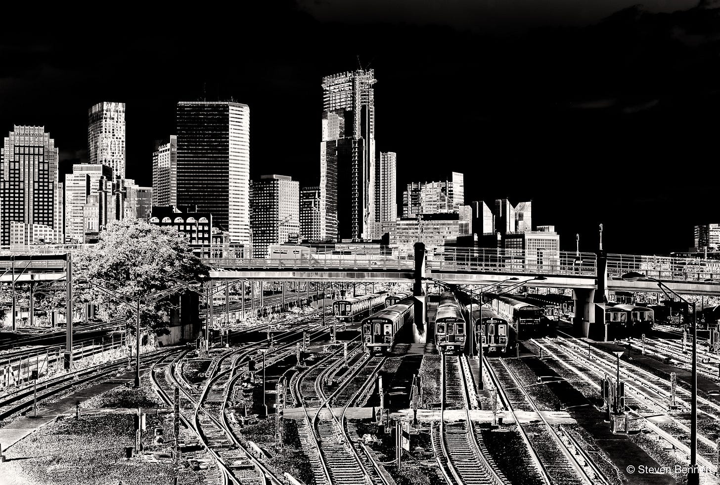 High-contrast black-and-white photo of Boston's downtown skyline and MBTA rail yard, viewed from the West 4th Street bridge, with a lone tree and crisscrossing train tracks in the foreground. High-contrast black-and-white photo of Boston's downtown skyline and MBTA rail yard, viewed from the West 4th Street bridge, with a lone tree and crisscrossing train tracks in the foreground.