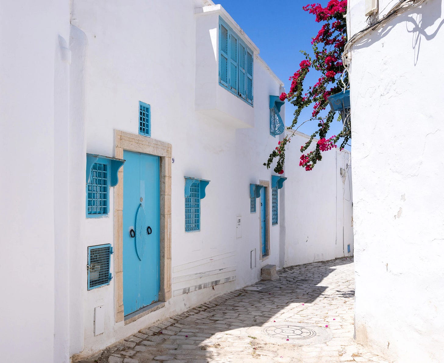 Sunlit alley in Sidi Bou Said, Tunisia, lined with whitewashed walls, turquoise doors, and windows accented by bright pink bougainvillea. The narrow cobblestone path captures the village's harmony of light, color, and scale that makes it feel both real and dreamlike.