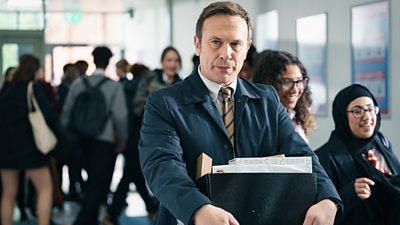 A man in a suit and dark blue jacket walks through a busy school corridor holding a box of things
