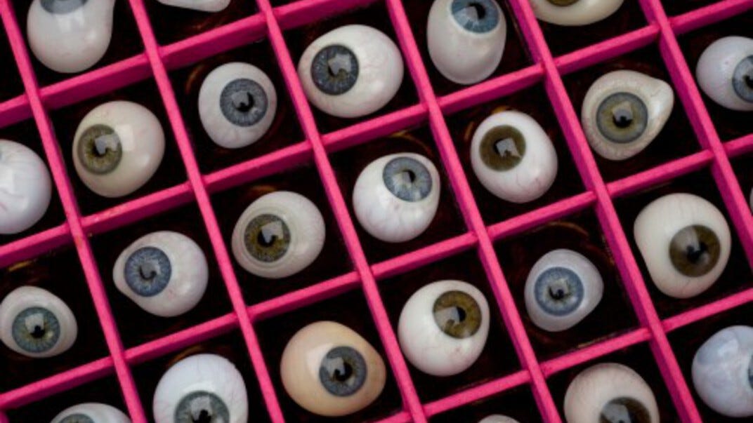 foto van een bak vol met kunstogen, in allerlei verschillende kleuren. Ieder oog ligt in een eigen vakje op een zwarte achtergrond, de vakjes zijn gemaakt van fel rozekleurig materiaal