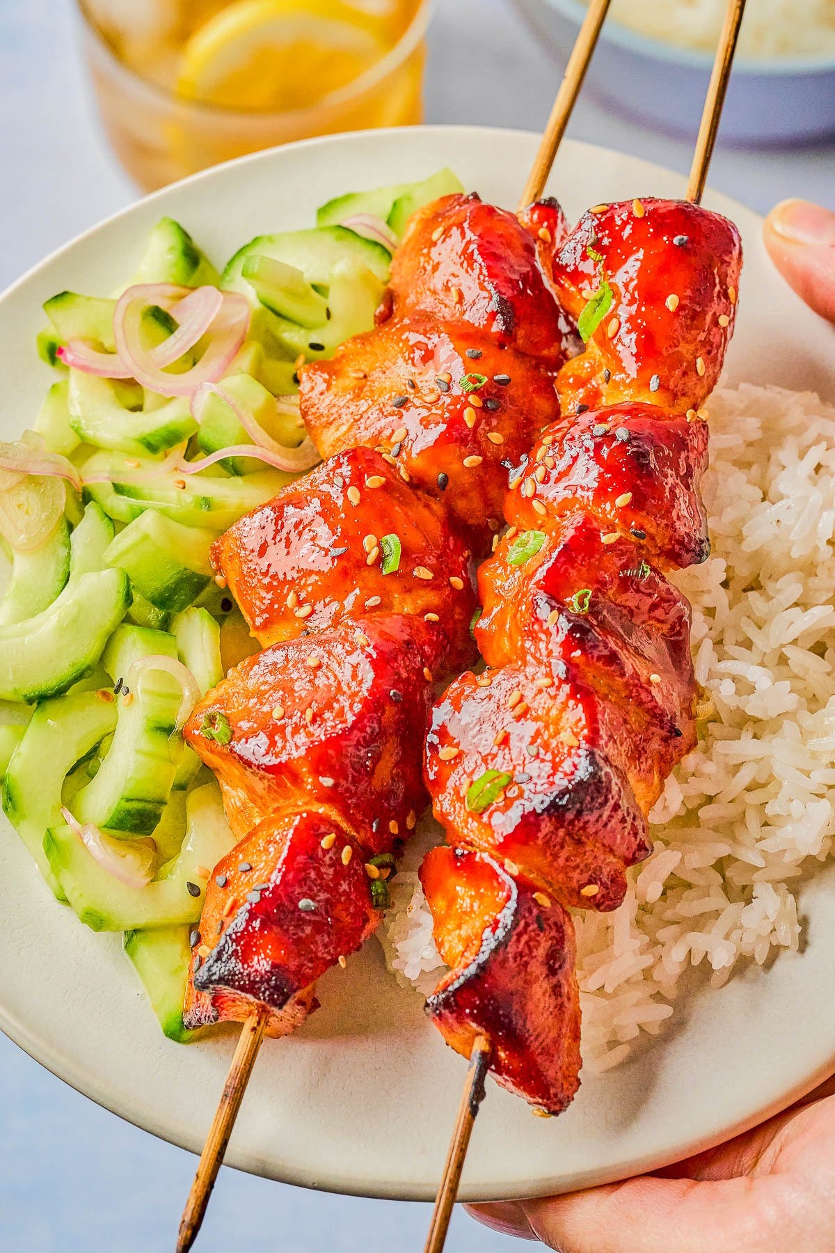 Two skewers of glazed grilled chicken served on white rice with a cucumber salad on a white plate.