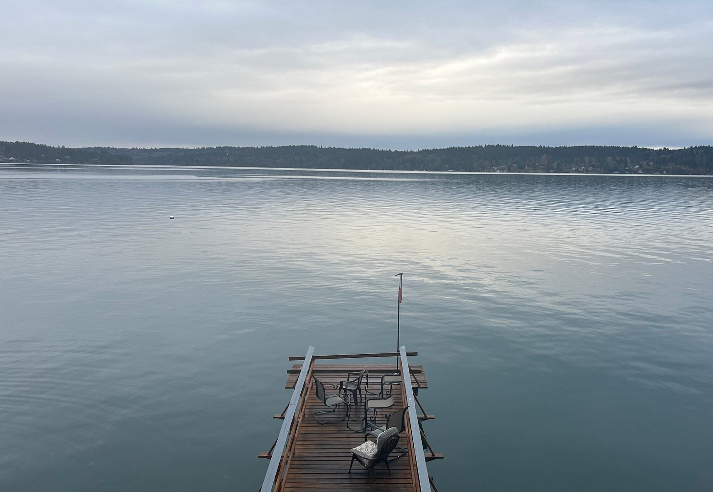 view of a small bit of wooden dock with five chairs extending out into the waters of the Puget Sound, with tree-covered hills in the distance. the water is slightly rippled and mirrors, in color and texture, the cloudy skies above. 