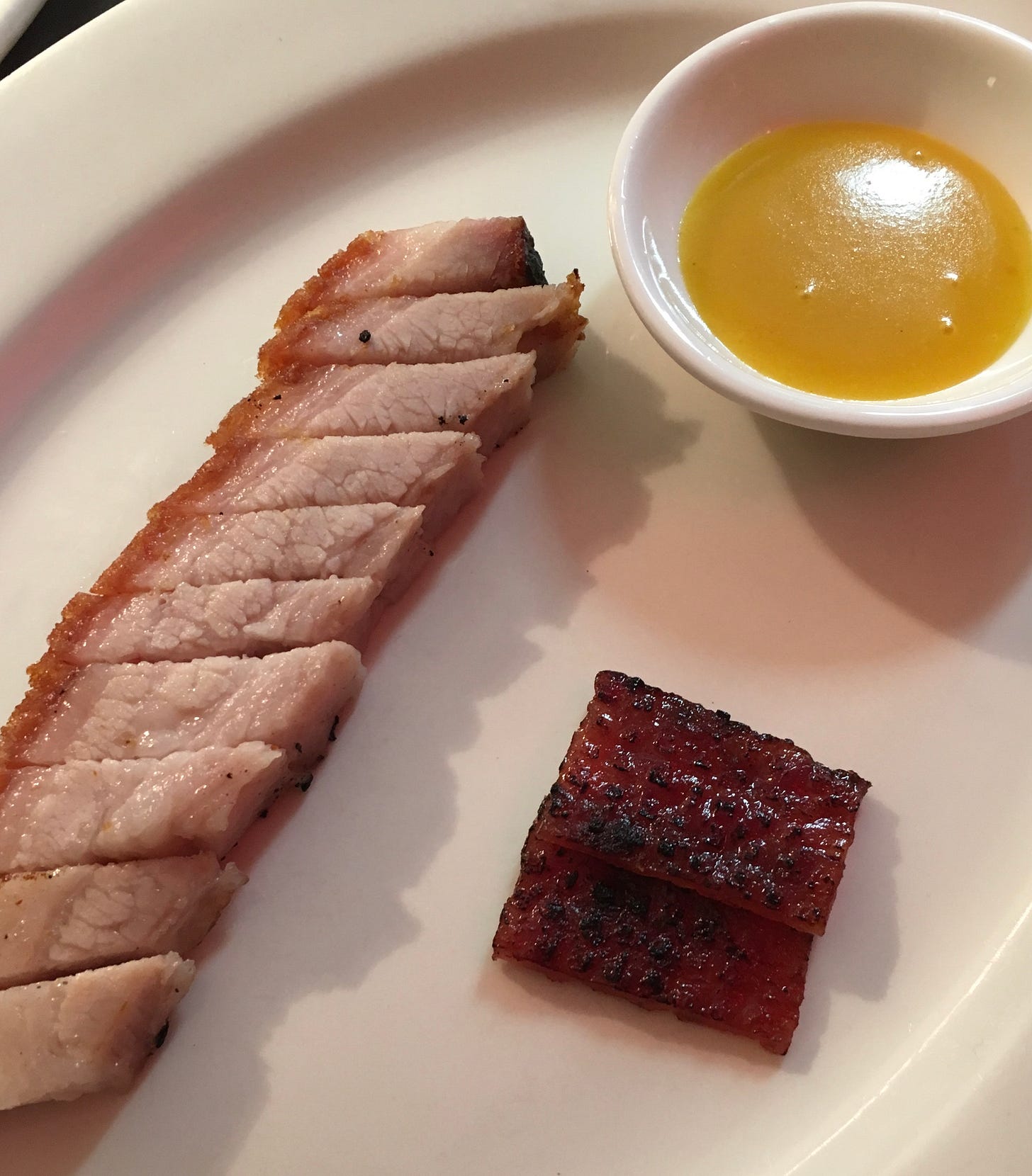 A thick cut of Cantonese crispy pork belly on its side on a white plate, with a serving of mustard sauce in a small white bowl and small portion of crispy belly skin, crisp-side up A thick cut of Cantonese crispy pork belly on its side on a white plate, with a serving of mustard sauce in a small white bowl and small portion of crispy belly skin, crisp-side up