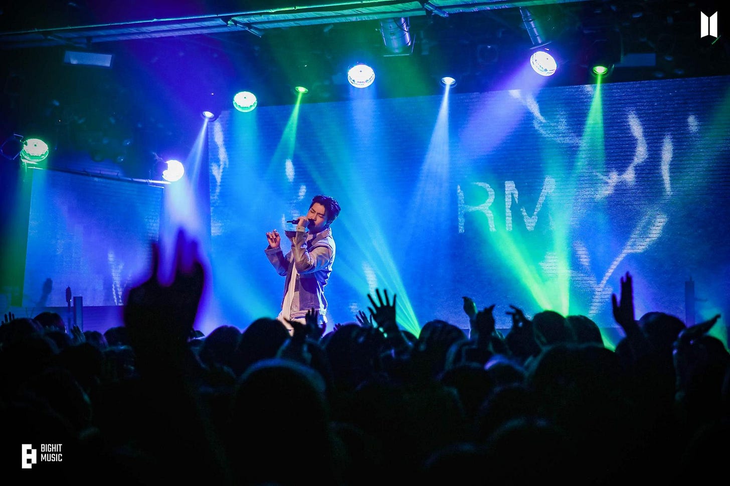 RM sings to a small crowd with blue lighting and "RM" projected on the screen behind him. RM sings to a small crowd with blue lighting and "RM" projected on the screen behind him.