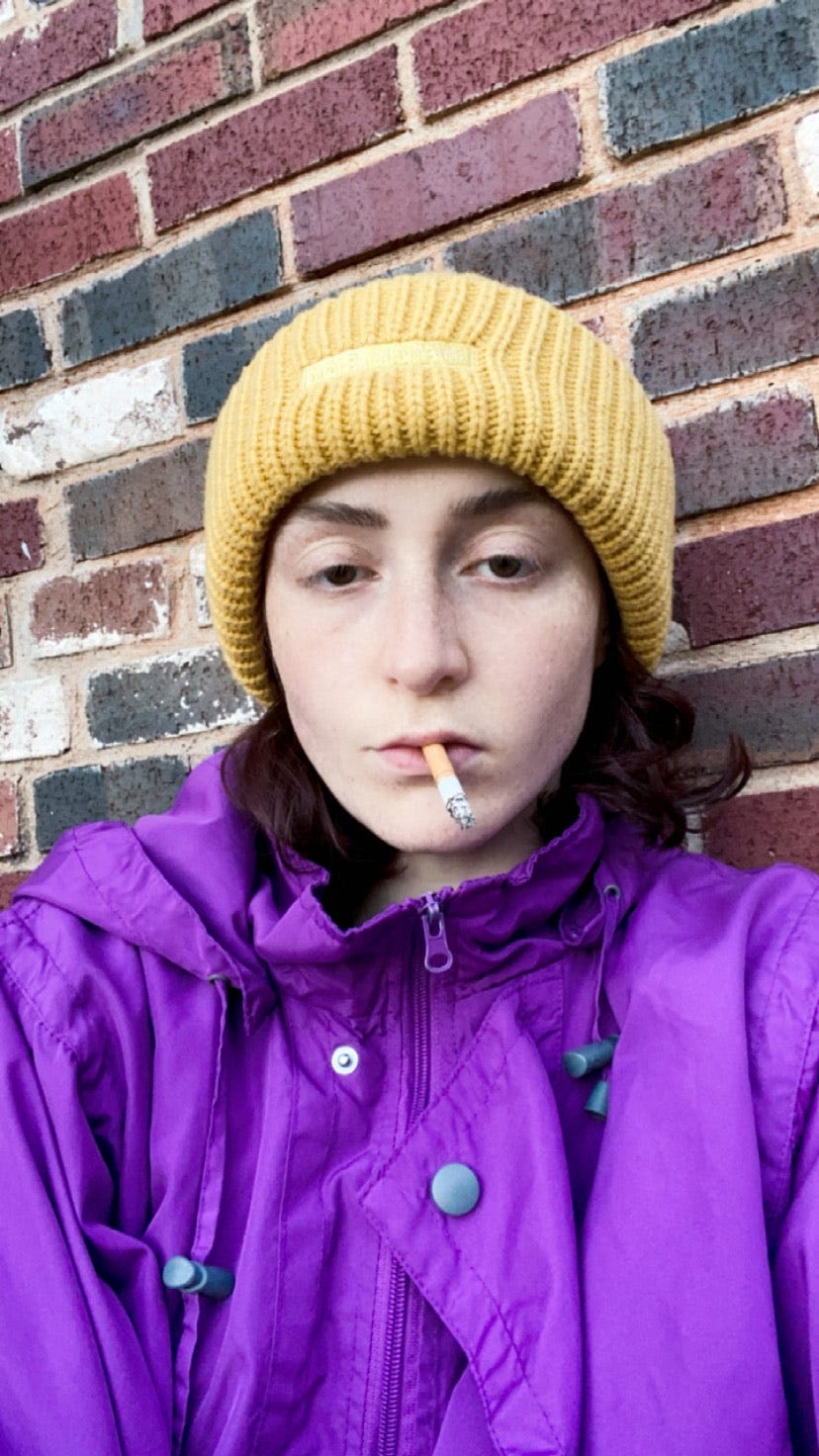 a photo of Jo in a purple rain jacket and yellow beanie smoking a cigarette in front of a brick wall