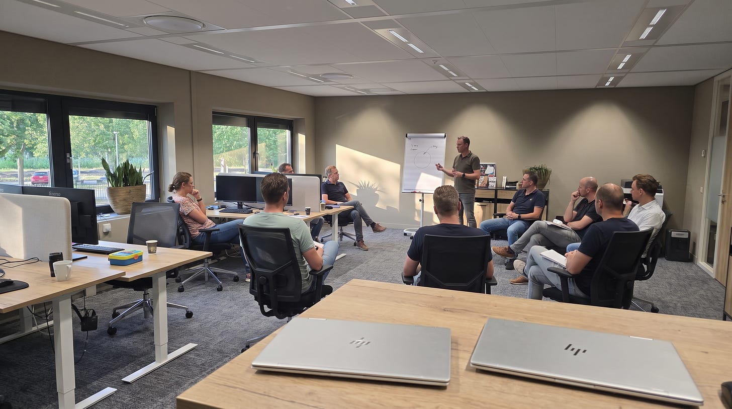 Op de werkvloer hebben collega's hun stoelen in een halve kring getrokken rond iemand die bij een flipover staat uit te leggen, met op de bureaus op de voorgrond twee dichtgeklapte laptops Op de werkvloer hebben collega's hun stoelen in een halve kring getrokken rond iemand die bij een flipover staat uit te leggen, met op de bureaus op de voorgrond twee dichtgeklapte laptops