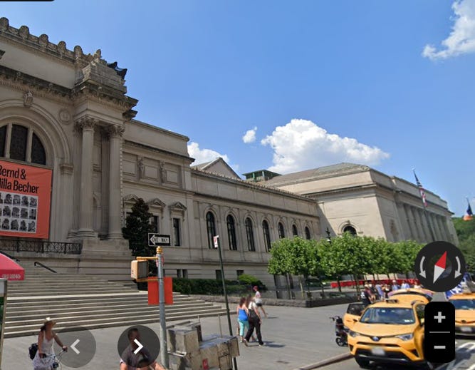 Screenshot of a partial view of a museum's entrance with cabs parked on the street and people on the sidewalk on a bright sunny day.