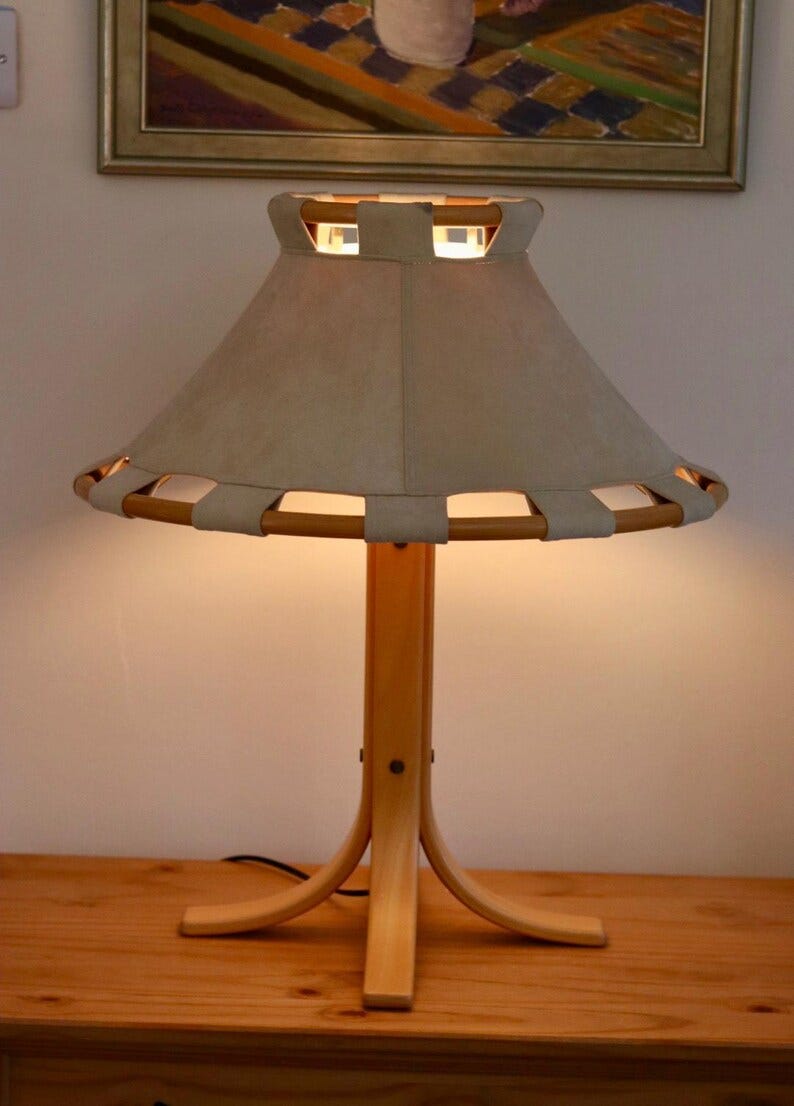 May include: A wooden table lamp with a light beige lampshade. The lamp has a light wood base with a curved design. The lampshade is a light beige color and has a unique design with cutouts. The lamp is turned on, emitting a warm glow. May include: A wooden table lamp with a light beige lampshade. The lamp has a light wood base with a curved design. The lampshade is a light beige color and has a unique design with cutouts. The lamp is turned on, emitting a warm glow.