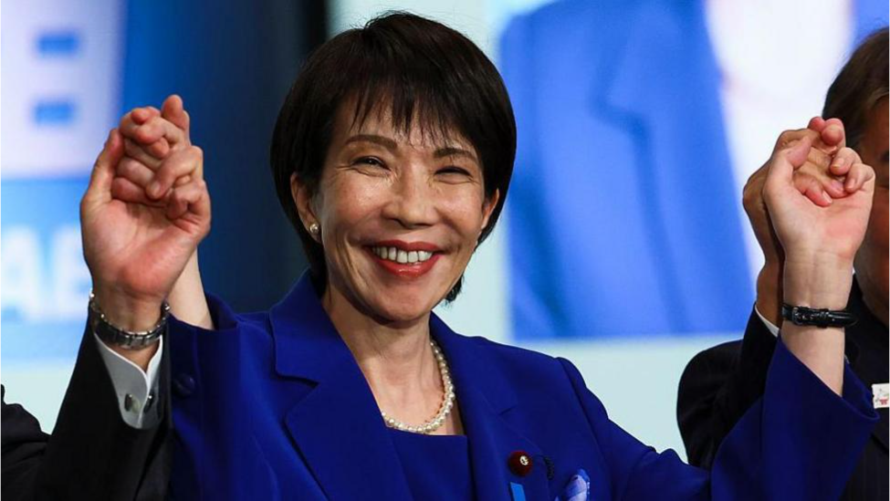 A woman with short black hair, wearing red lipstick and a blue suit and pears, smiles widely. She is holding the hands of two people on either side of her, and they raise their hans in the air.