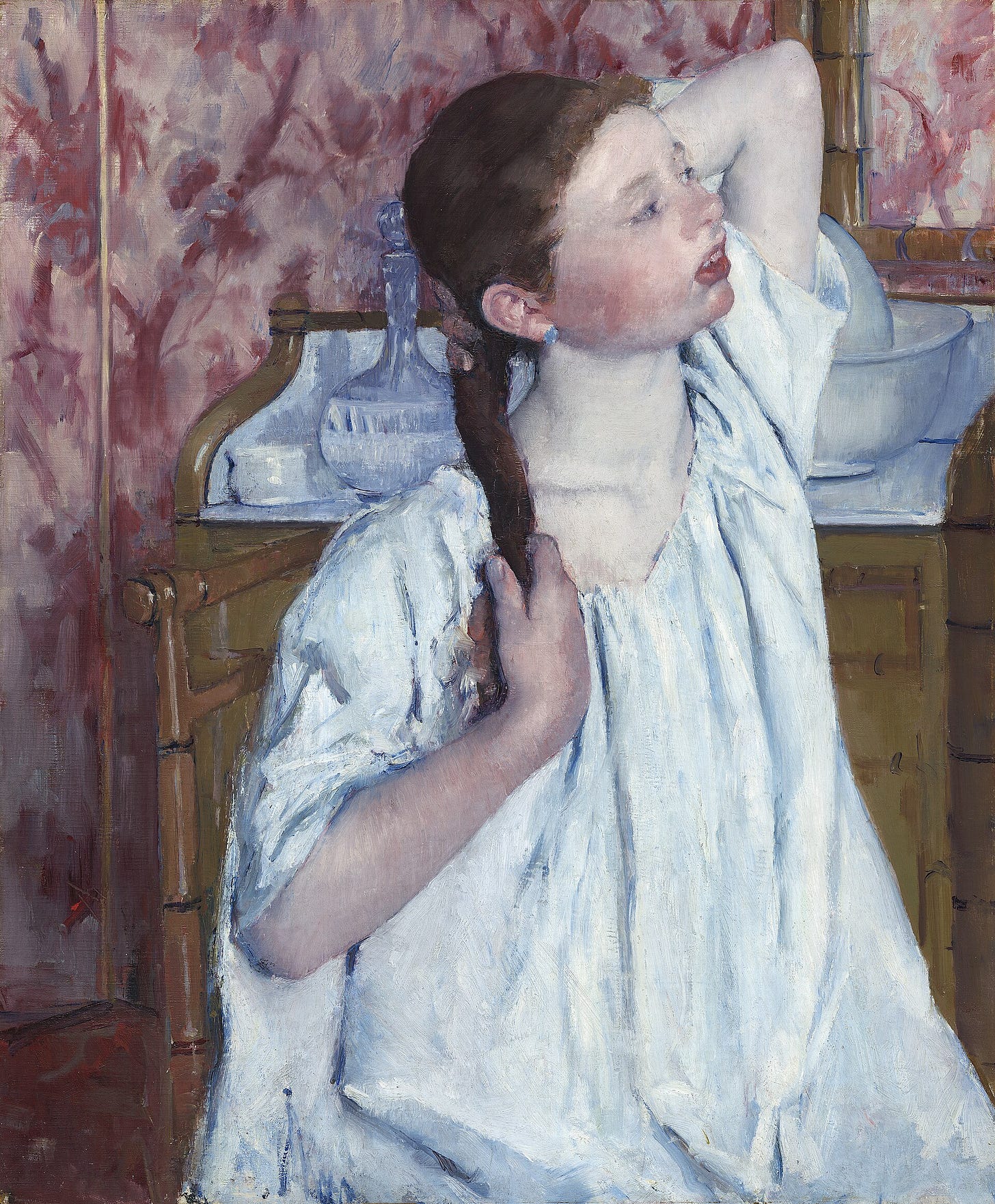 A young woman in a loose white chemise sits on a low sofa, holding her long brown hair with both hands at the back of her neck.
