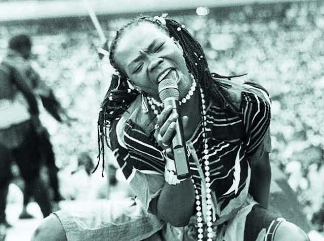 A black and white photo of a woman singinging her heart out into a microphone on stage, her braided hair in pigtails, her eyes shut. She's down on her haunches. A black and white photo of a woman singinging her heart out into a microphone on stage, her braided hair in pigtails, her eyes shut. She's down on her haunches.