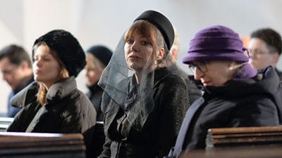 Philomena Cunk (Diane Morgan) sitting in a church dressed in black with a black veil over her face and looking to the camera