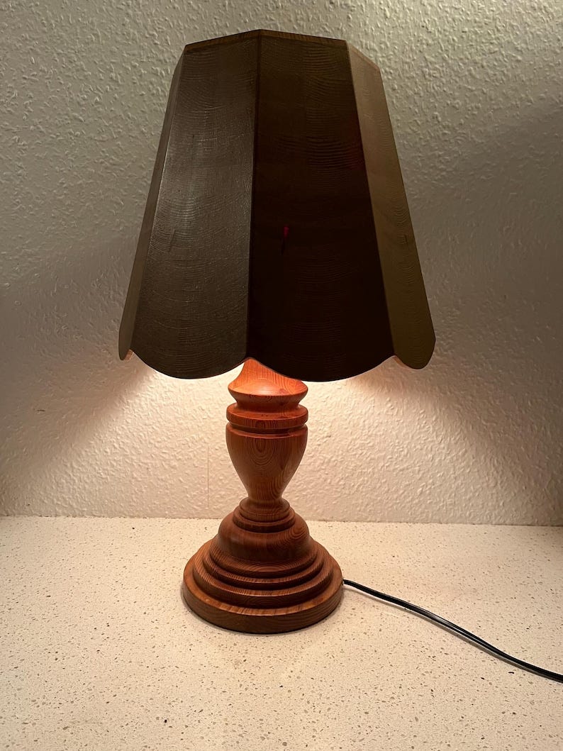 May include: A wooden table lamp with a dark brown, scalloped lampshade. The lamp base is a rich brown color and has a turned, fluted design. May include: A wooden table lamp with a dark brown, scalloped lampshade. The lamp base is a rich brown color and has a turned, fluted design.