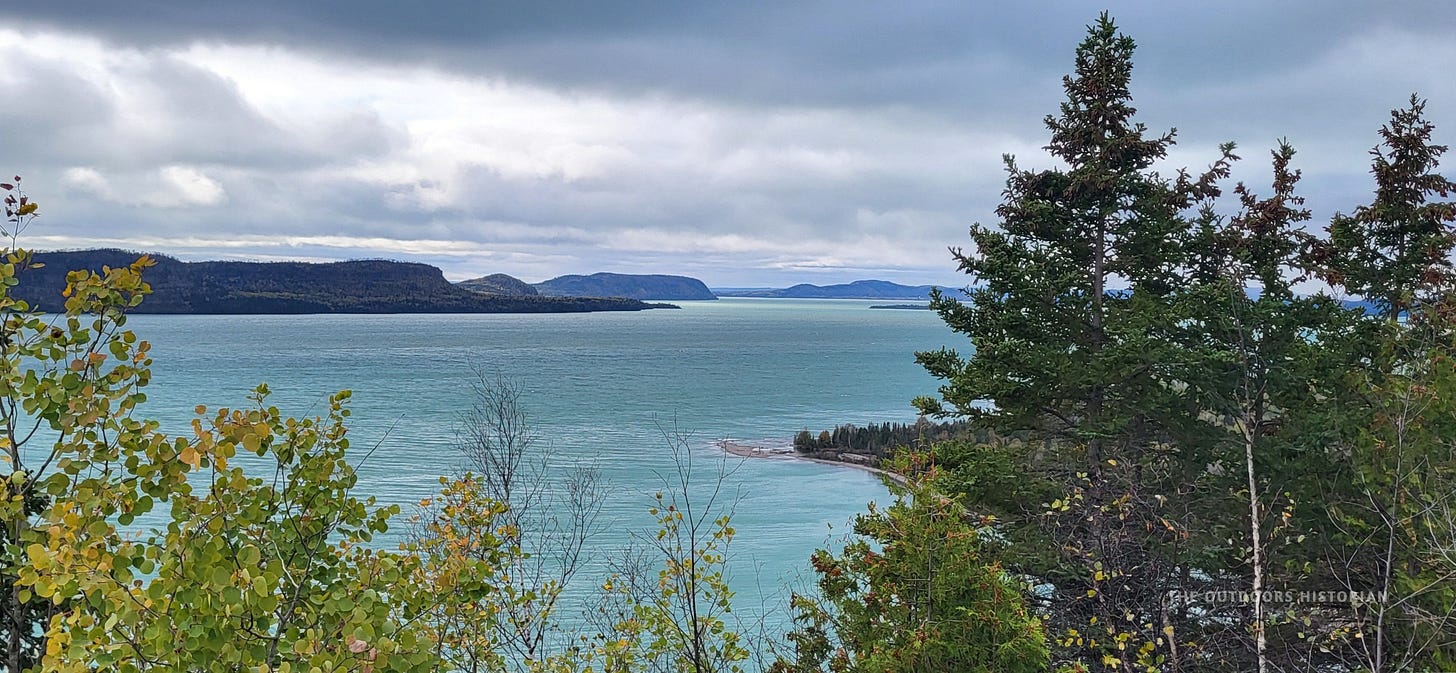 A high-elevation view looking down at Lake Superior. The water in the bay below is a striking, bright turquoise blue, contrasting with the dark evergreen forest and grey rocks in the foreground.