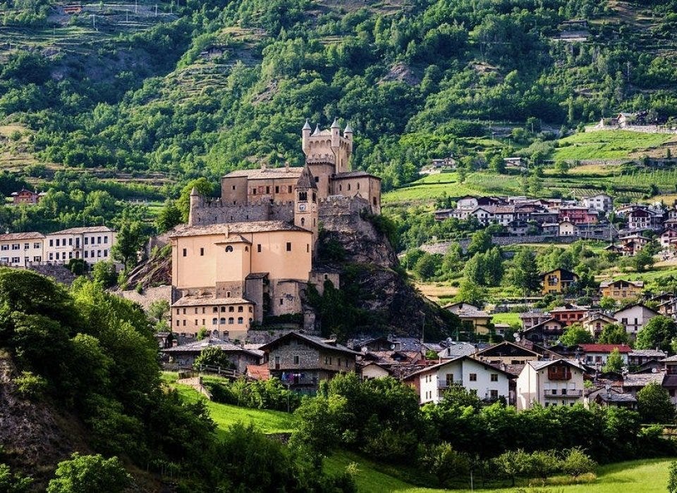 The medieval Saint-Pierre Castle and its parish church built on a rocky spur, overlooking an alpine village on a green hillside in northern Italy. The medieval Saint-Pierre Castle and its parish church built on a rocky spur, overlooking an alpine village on a green hillside in northern Italy.