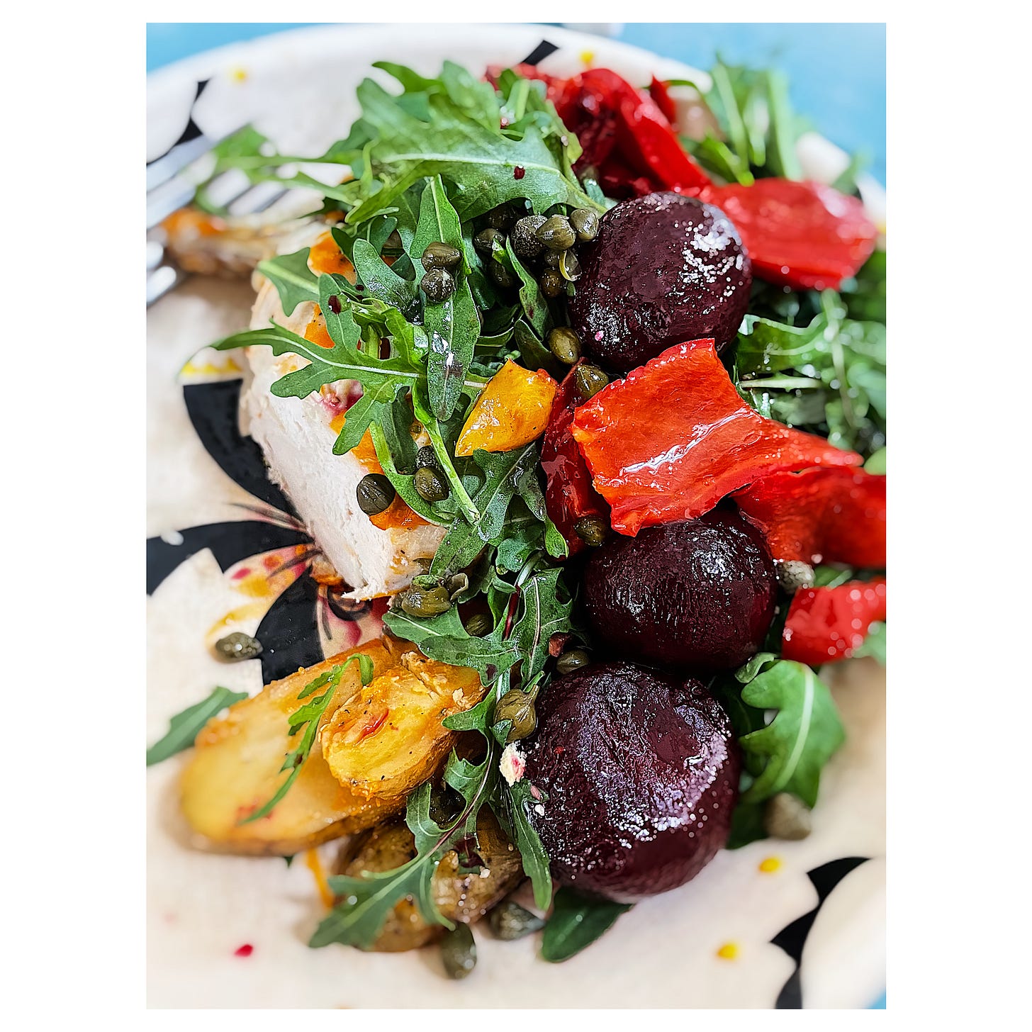A close up arial photograph of a bowl of salad consisting of rocket leaves, capers, roasted peppers, capers, roasted new potatoes and a mostly hidden chicken breast. Part of the ceramic bowl is visible, it is cream coloured with a black and yellow pattern. The prongs of a fork can be seen in the back right corner of the picture.