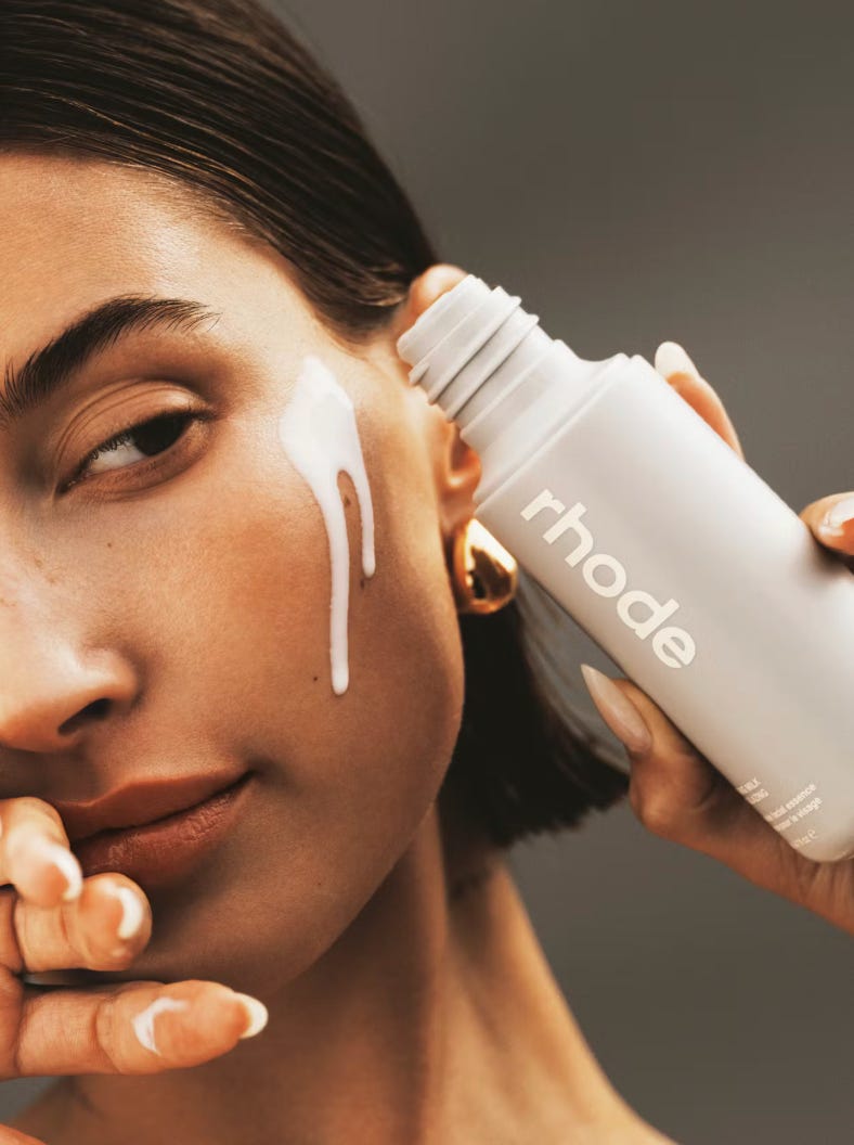 A close-up, high-definition photograph of Hailey Bieber’s face, highlighting a dewy, hydrated skin texture. She is holding a gray bottle of Rhode Glazing Milk near her cheek. The focus is on the "glazed" finish of her skin and the minimalist brand packaging. A close-up, high-definition photograph of Hailey Bieber’s face, highlighting a dewy, hydrated skin texture. She is holding a gray bottle of Rhode Glazing Milk near her cheek. The focus is on the "glazed" finish of her skin and the minimalist brand packaging.