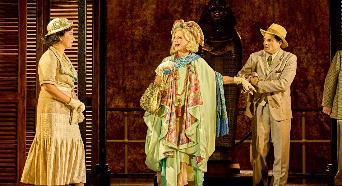 Three people on a stage wearing gold, blue and teal costumes, with a dark wooden backdrop behind them Three people on a stage wearing gold, blue and teal costumes, with a dark wooden backdrop behind them