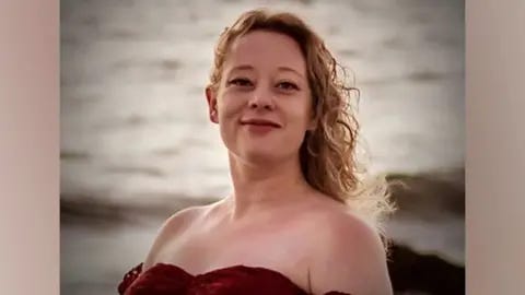 Renee Good has long, blonde, curly hair and is wearing a red off-the-shoulder top. The ocean is in the background.