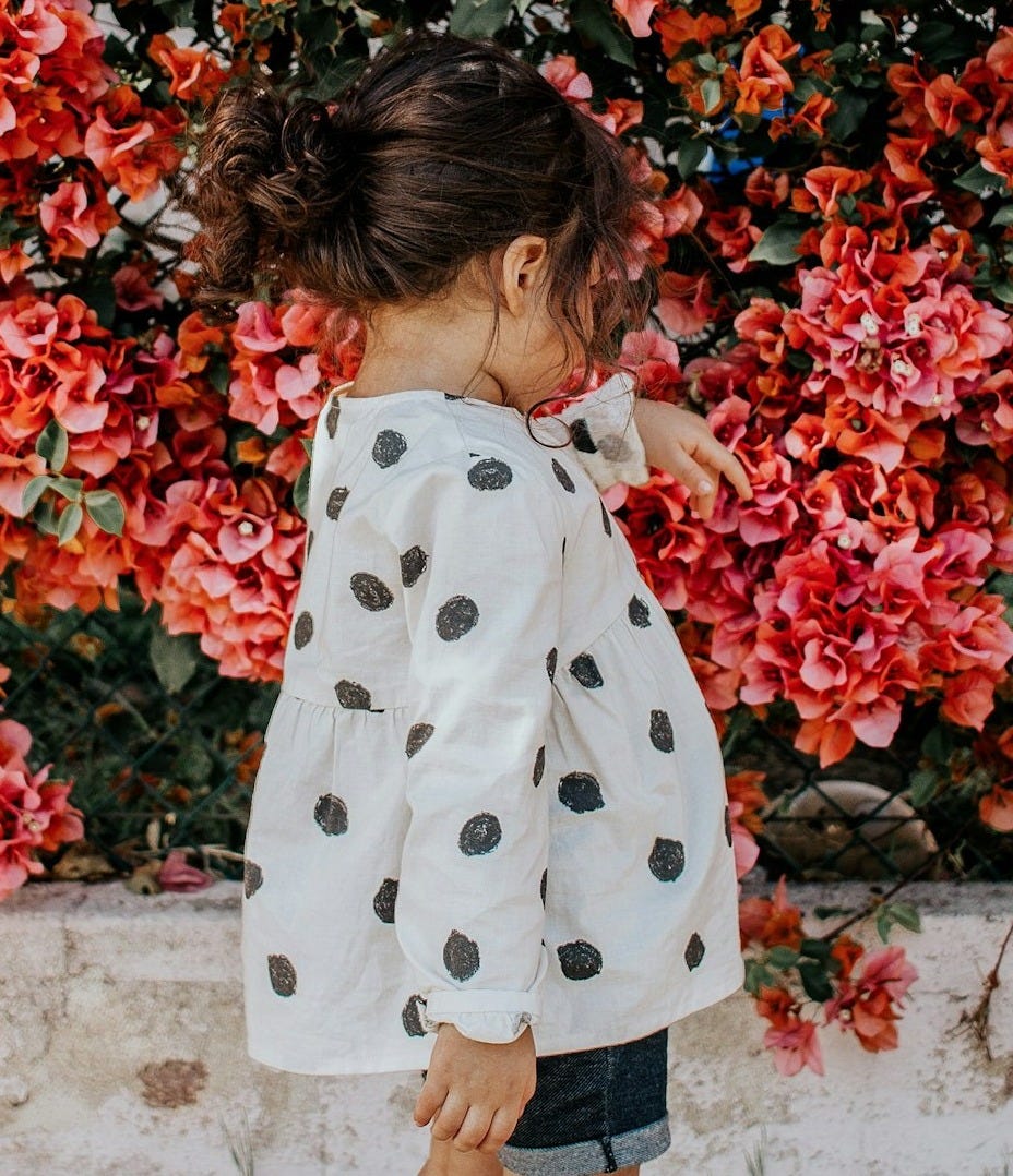 girl in black and white polka-dot long-sleeved blouse and blue shorts standing beside red petaled flowers during daytime