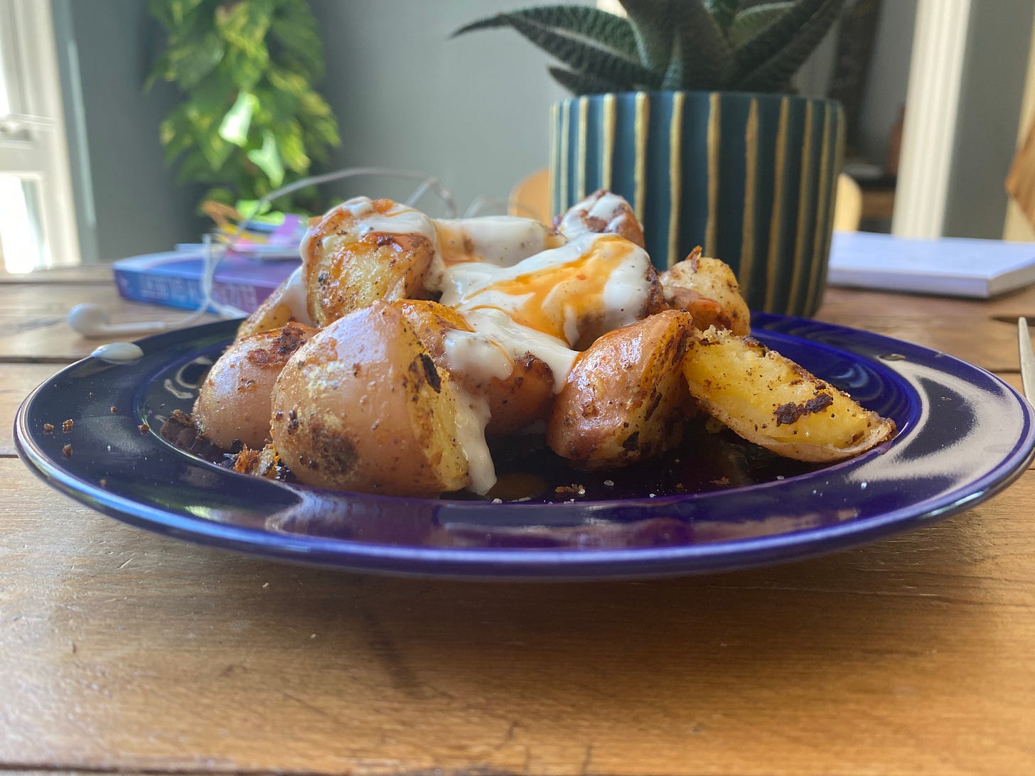 Blue plate topped with fried potatoes and sauces. The plate is on a table. A pot plant, books and headphones are in the background.