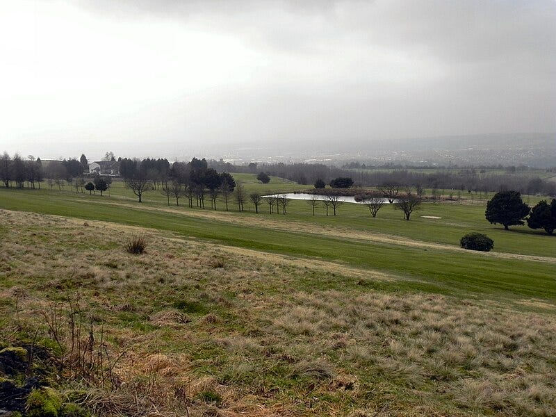 A golf course on an overcast day. Trees disappear into the fog further downhill. A building, pond, and houses in the distance are barely visible.