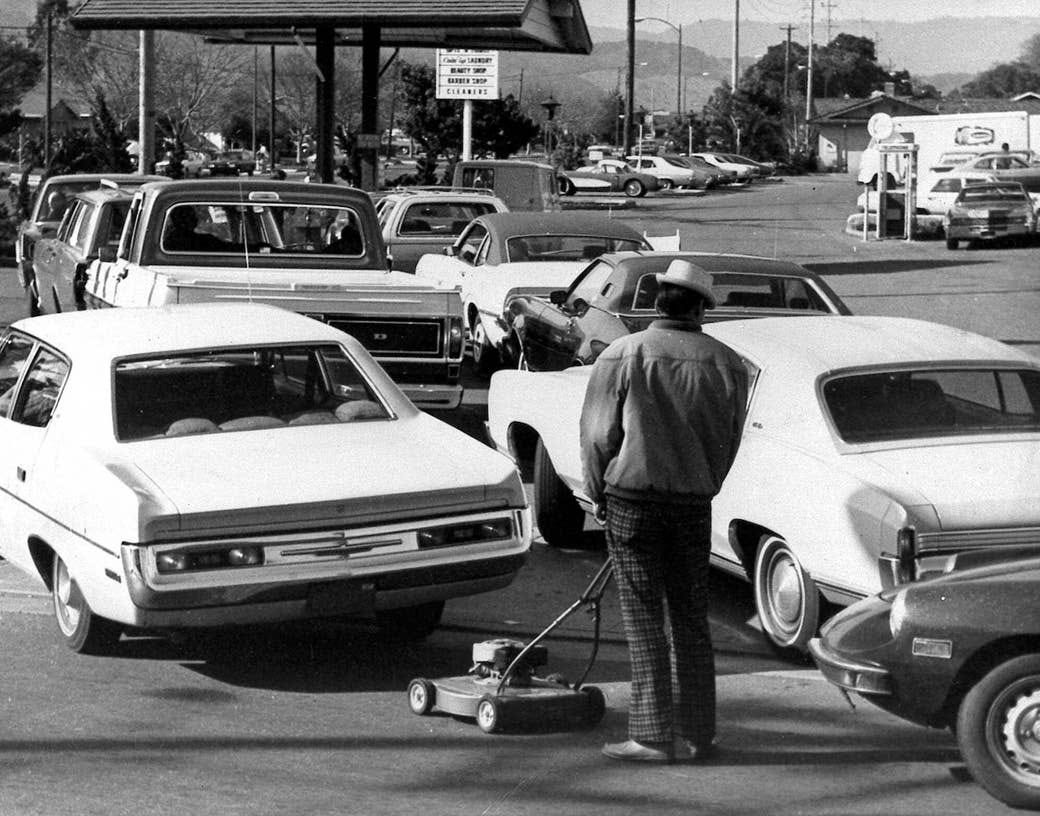 A person standing behind a line of cars pushes his lawnmower