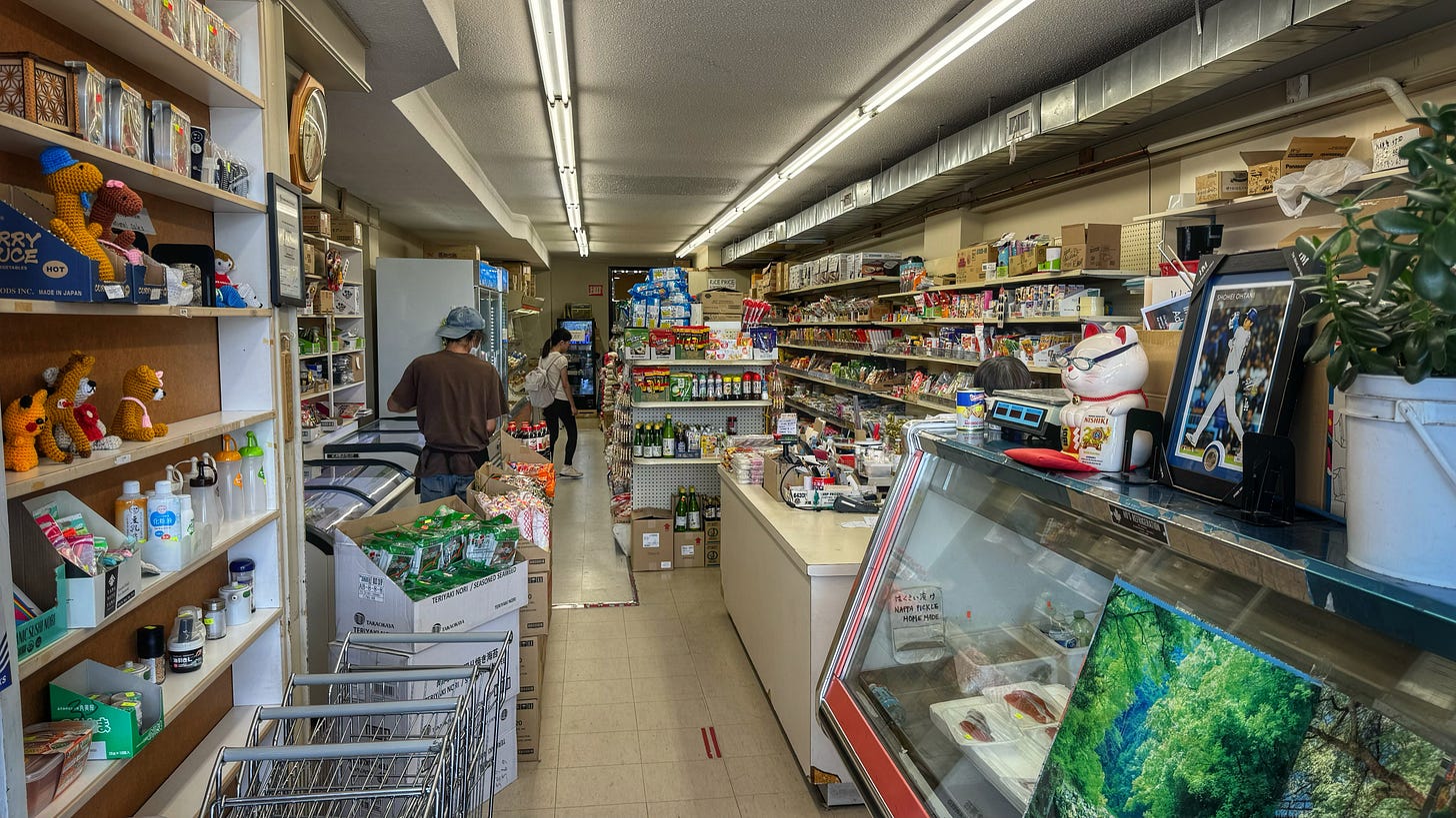 A photo taken from just inside the front door of Sandown Market. On the left side there is a series of shelves, fridges, and freezers. On the right there are a refrigerated display case, a cash register, and, farther back, more shelves. Everything is very plain and slightly cluttered.