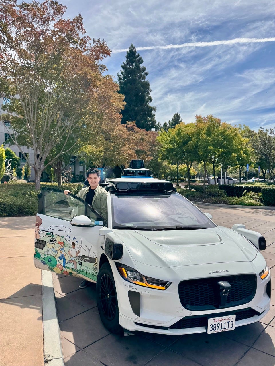 The author smiling behind the open door of a white Waymo Jaguar I-PACE robotaxi parked on a sunny, tree-lined plaza at Waymo HQ, its rooftop lidar dome and sensor pods visible and a colorful mural painted on the open door.