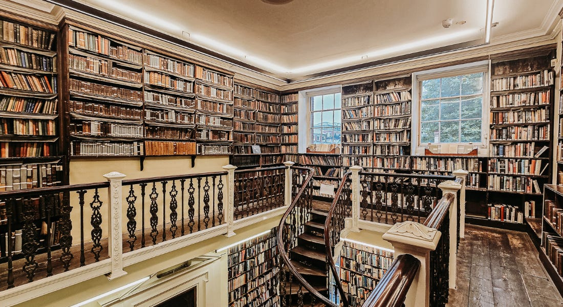 A library with brown bookshelves, colourful books, white walls, two windows, and a brown spiral staircase A library with brown bookshelves, colourful books, white walls, two windows, and a brown spiral staircase