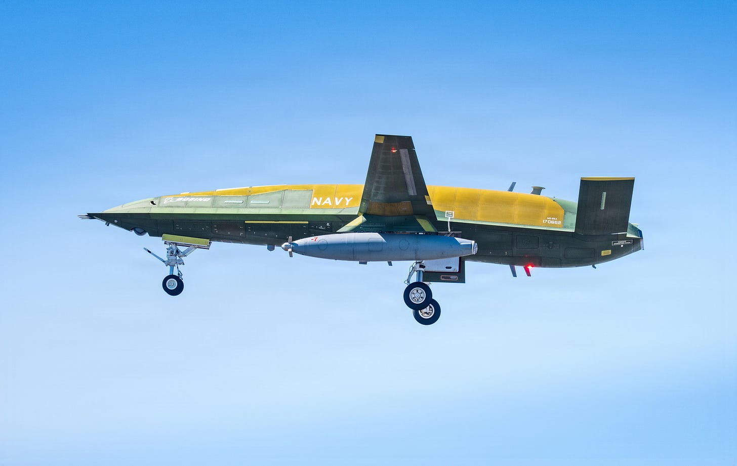 A Boeing MQ-25A Stingray in level flight against a clear blue sky, seen from the side with its landing gear down. The aircraft has no cockpit and no windows. Its fuselage is finished in unpainted green and yellow primer, marked NAVY and numbered 170662, with a large gray external pod slung beneath it.