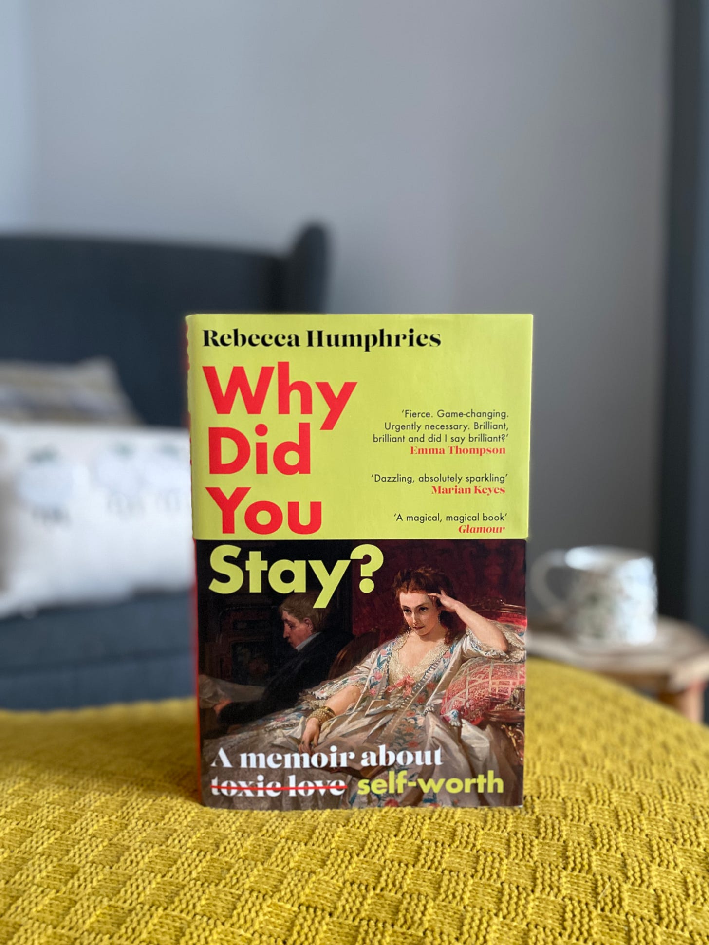 A book, Why Did You Stay, sits on a footstool in front of an armchair that's blurred in the background.