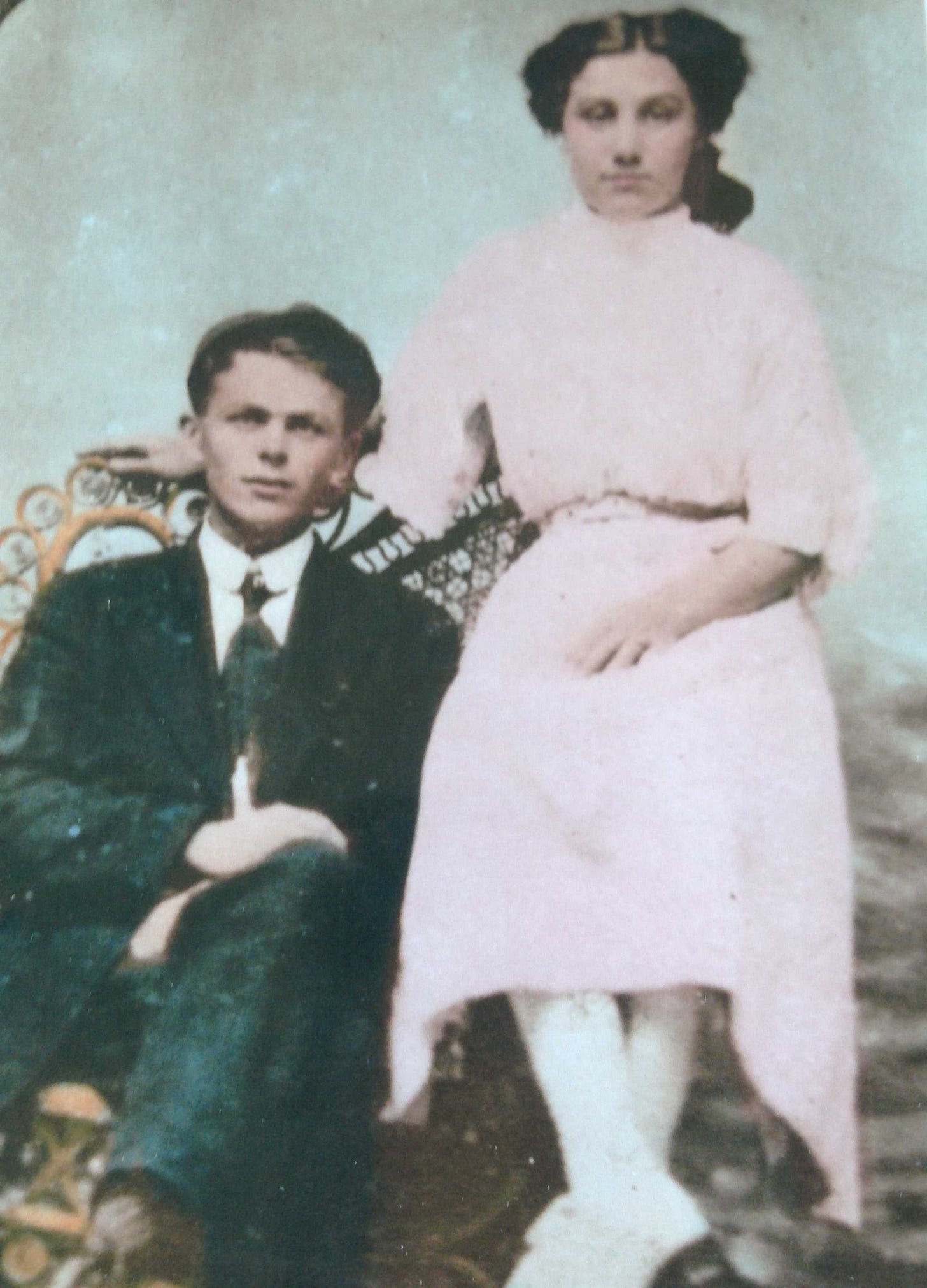 A photograph, apparently hand-tinted, of a young couple in the years immediately after the turn of the 19th to 20th centuries. The man is dressed neatly in a suit and tie, and seated in a large wicker chair. The woman wears a light-colored (pink?) dress, buttoned to the chin, and white hosiery; she sits on the left arm of the wicker chair, and her right arm is draped across the back. They seem very content with their lot in life. A photograph, apparently hand-tinted, of a young couple in the years immediately after the turn of the 19th to 20th centuries. The man is dressed neatly in a suit and tie, and seated in a large wicker chair. The woman wears a light-colored (pink?) dress, buttoned to the chin, and white hosiery; she sits on the left arm of the wicker chair, and her right arm is draped across the back. They seem very content with their lot in life.