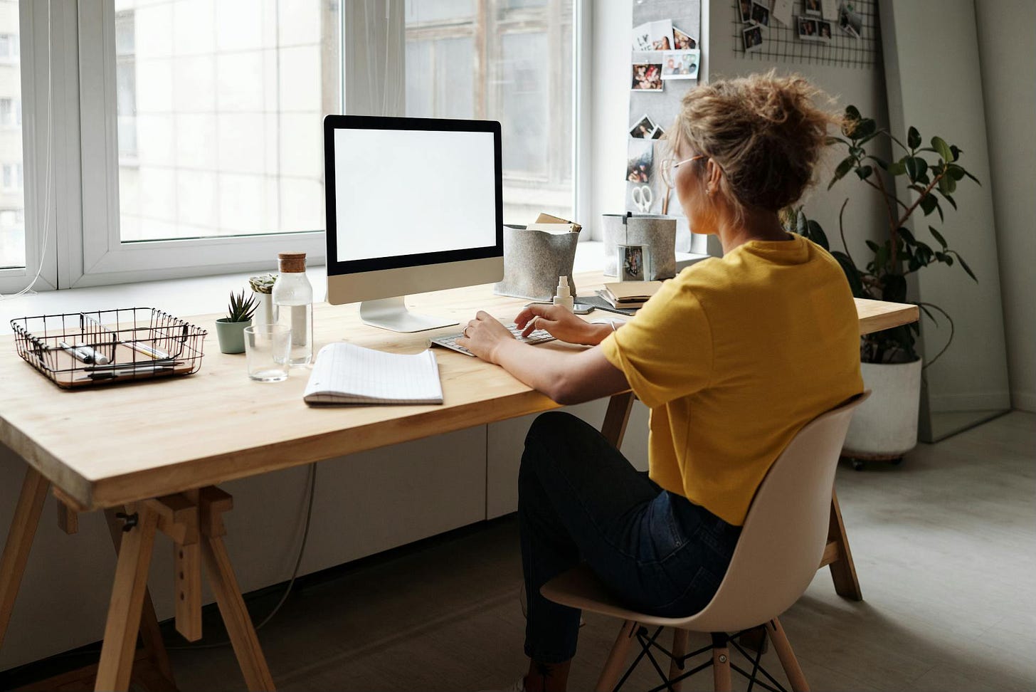 A woman in a mustard-colored top and black pants works on a computer sitting on a large desk in front of a window.