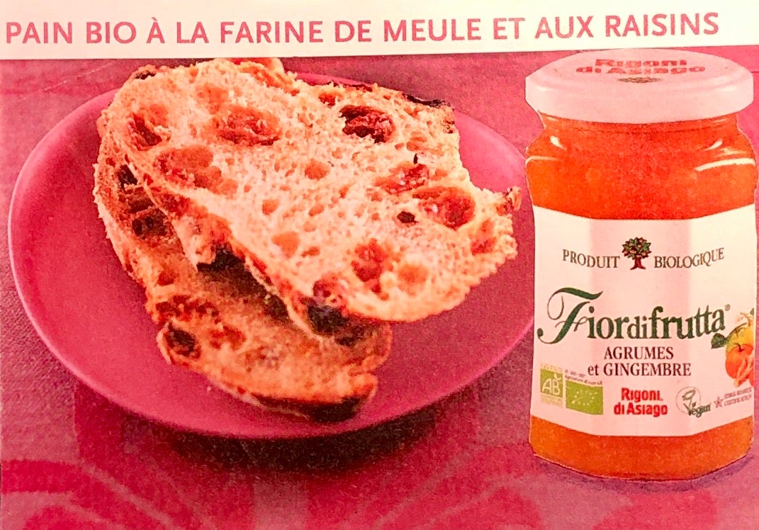 Une nappe rose à motifs rouges sur laquelle est posée une petite assiette rose contenant des tranches de pain aux fruits secs. Au premier plan : un pot de gelée d'agrumes. En haut de l'image est collé le texte "pain bio à la farine de meule et aux raisins".
