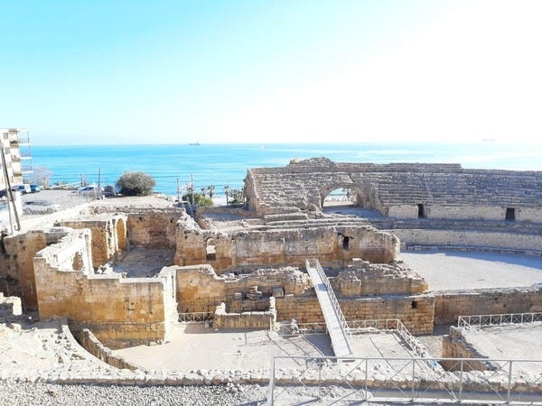 Ruínas de um Anfiteatro Romano em Tarragona. É possível ver as arquibancadas do anfiteatro. Dentro, paredes que correspondiam a construções cristãs de muitos anos depois da derrocada do anfiteatro. Perto da ponte, na parte inferior da foto, é possível ver uma lápide escavada do cemitério que existia muito tempo antes do anfiteatro. Foto de Edmara Galvão. Descrição da Imagem: Ao fundo, o mesmo céu azul e sem nuvens e o mar calmo com tons azuis da imagem anterior. Em primeiro plano, ruínas de um anfiteatro romano. São arquibancadas de pedra, já bem gastas. No centro do anfiteatro, paredes erguidas com outro tipo de pedra, mais retangular, que formam as ruínas de uma construção antiga. Grades de ferro cercam várias partes do caminho. Ruínas de um Anfiteatro Romano em Tarragona. É possível ver as arquibancadas do anfiteatro. Dentro, paredes que correspondiam a construções cristãs de muitos anos depois da derrocada do anfiteatro. Perto da ponte, na parte inferior da foto, é possível ver uma lápide escavada do cemitério que existia muito tempo antes do anfiteatro. Foto de Edmara Galvão. Descrição da Imagem: Ao fundo, o mesmo céu azul e sem nuvens e o mar calmo com tons azuis da imagem anterior. Em primeiro plano, ruínas de um anfiteatro romano. São arquibancadas de pedra, já bem gastas. No centro do anfiteatro, paredes erguidas com outro tipo de pedra, mais retangular, que formam as ruínas de uma construção antiga. Grades de ferro cercam várias partes do caminho.