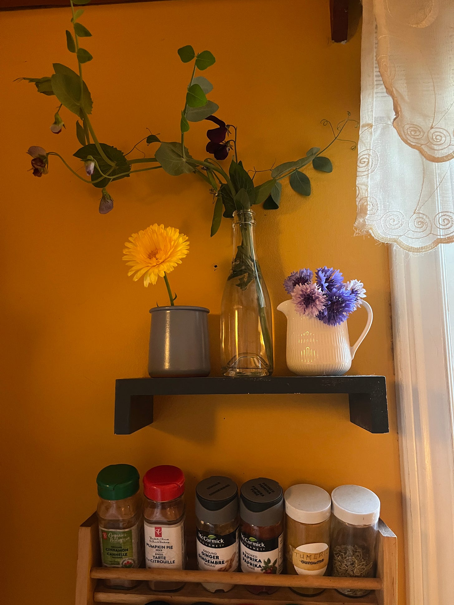 A small shelf in my kitchen above the spice rack with three little vases containing flowers.