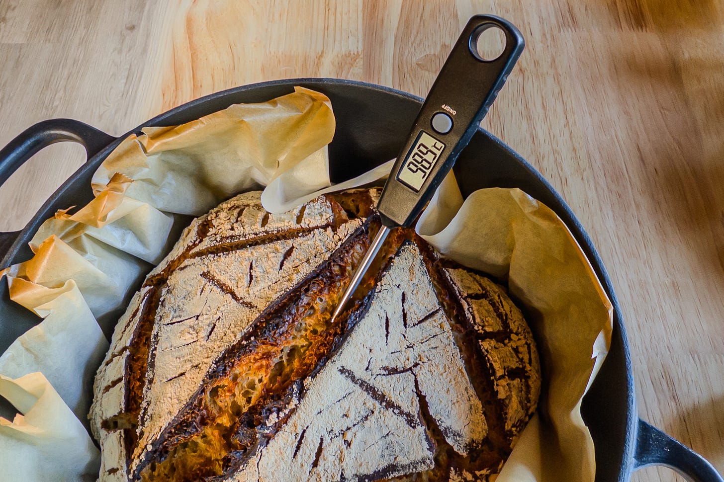 Draufsicht auf ein selbst gebackenes Sauerteigbrot direkt nach dem Backen. Es liegt in einem schwarzen Gusseisen-Topf auf Backpapier, in der Mitte steckt ein Digitalthermometer mit 98,9 Grad. Das Brot aus Hartweizenmehl sowie einer aromatischen Mischung aus Vollkornweizen-, Gerstenmalz- und geröstetem Roggenmehl zeigt eine rustikal aufgerissene, dunkel geröstete Kruste und eine hell bemehlte, dekorativ eingeschnittene Oberfläche.