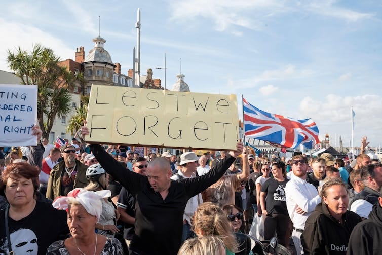 A man at a protest holding up a sign reading 'lest we forget'.